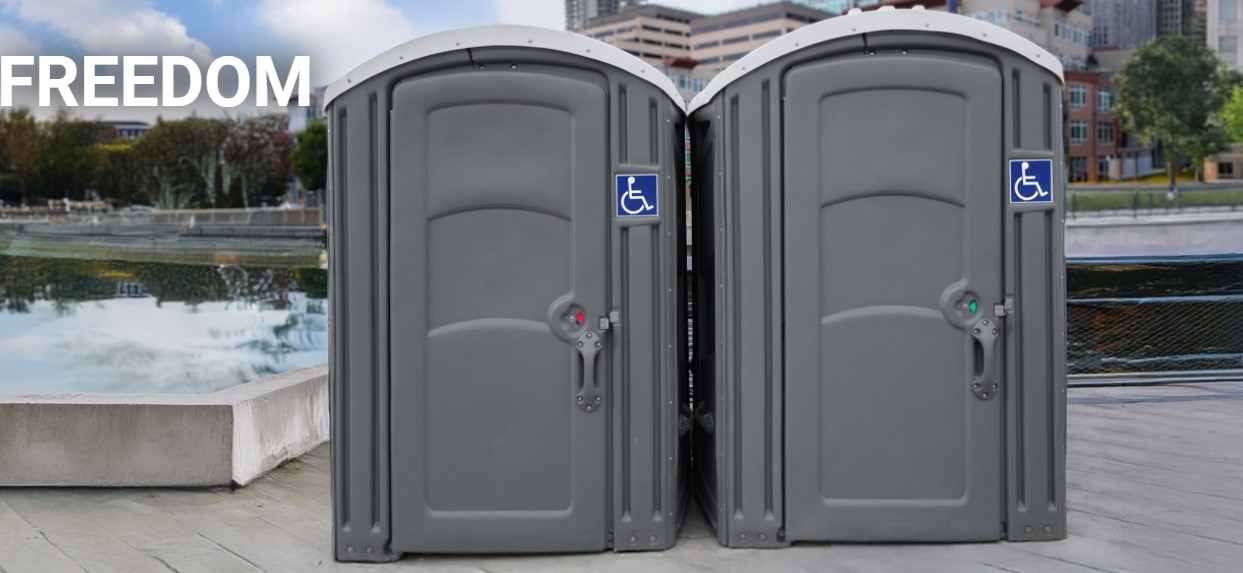 Two grey ADA compliant portable toilets near a pond in a city.